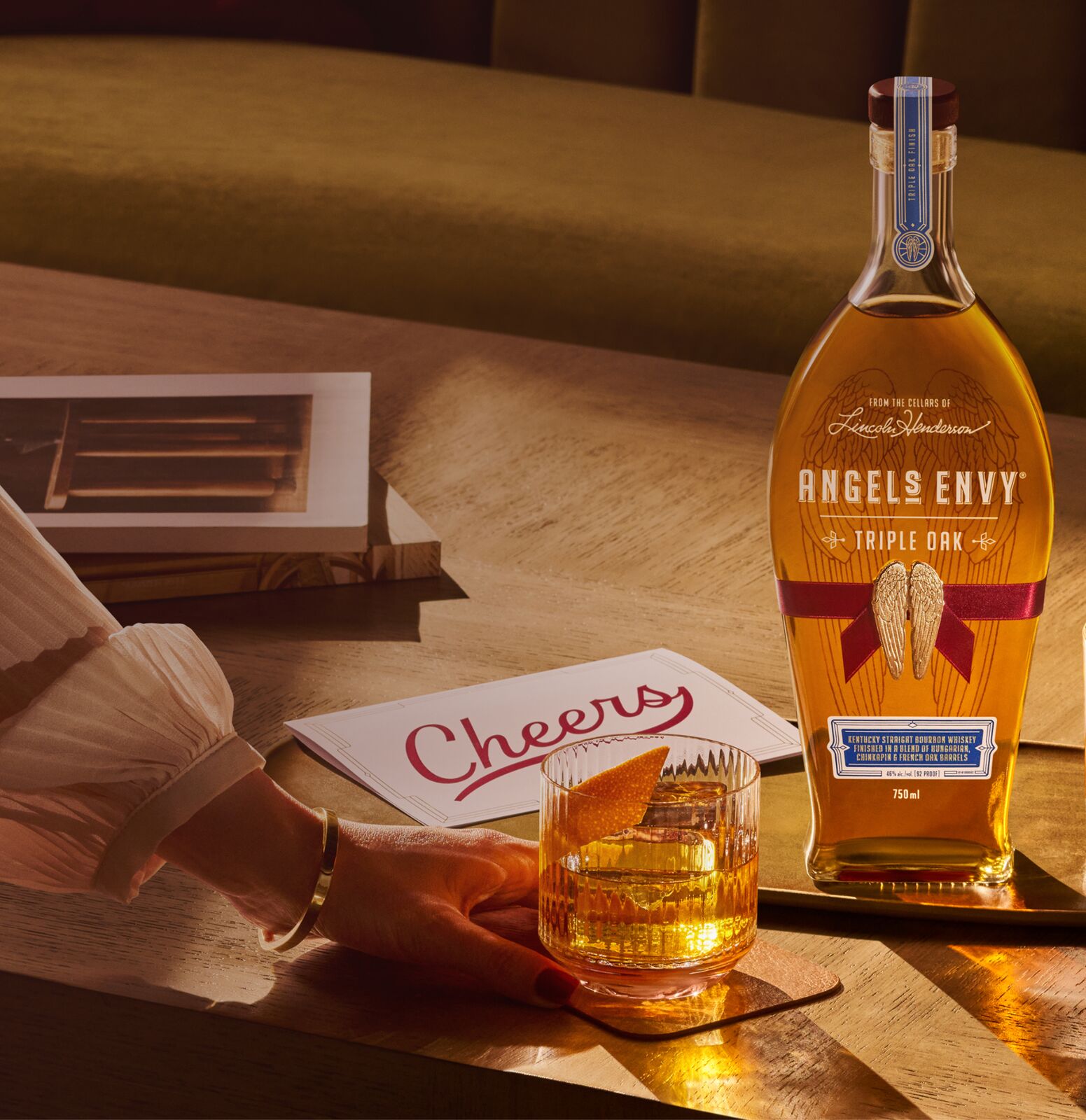 A bottle of Angel’s Envy Triple Oak bourbon decorated with a ribbon, sitting beside a handwritten “Cheers” card. A woman’s hand is reaching out to pick up an old fashioned cocktail.