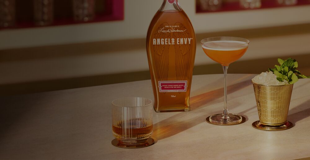 A sunlit bar featuring a bottle of Angel’s Envy bourbon presented with three polished drinks: bourbon served neat, a paper plane, and a mint julep.