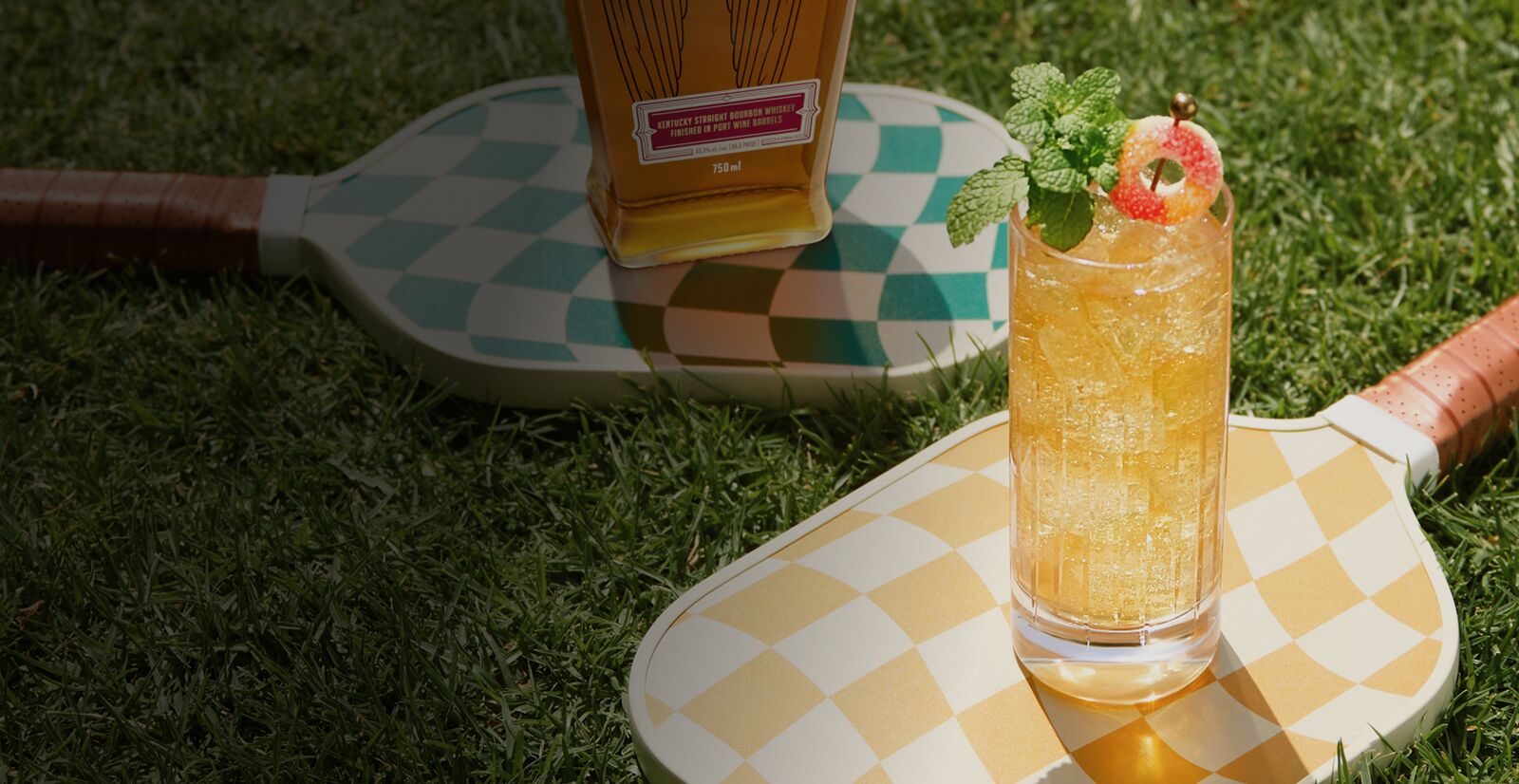 An icy Angel’s Envy Peach Smash cocktail garnished with mint and sour candy. It’s served in a highball glass on top of a patterned, pickleball paddle with a bottle of Angel’s Envy bourbon in the grassy background.