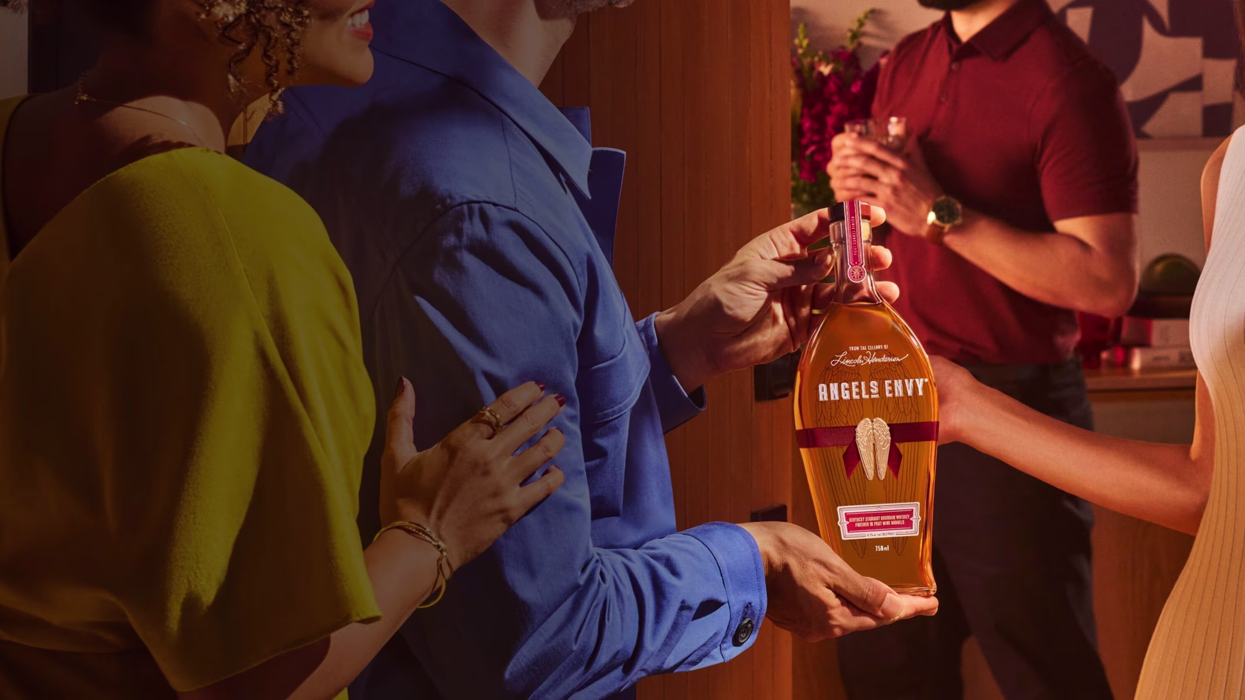 A guest gifting a bottle of Angel’s Envy bourbon to the host of a warm, stylish gathering, with friends in the background.