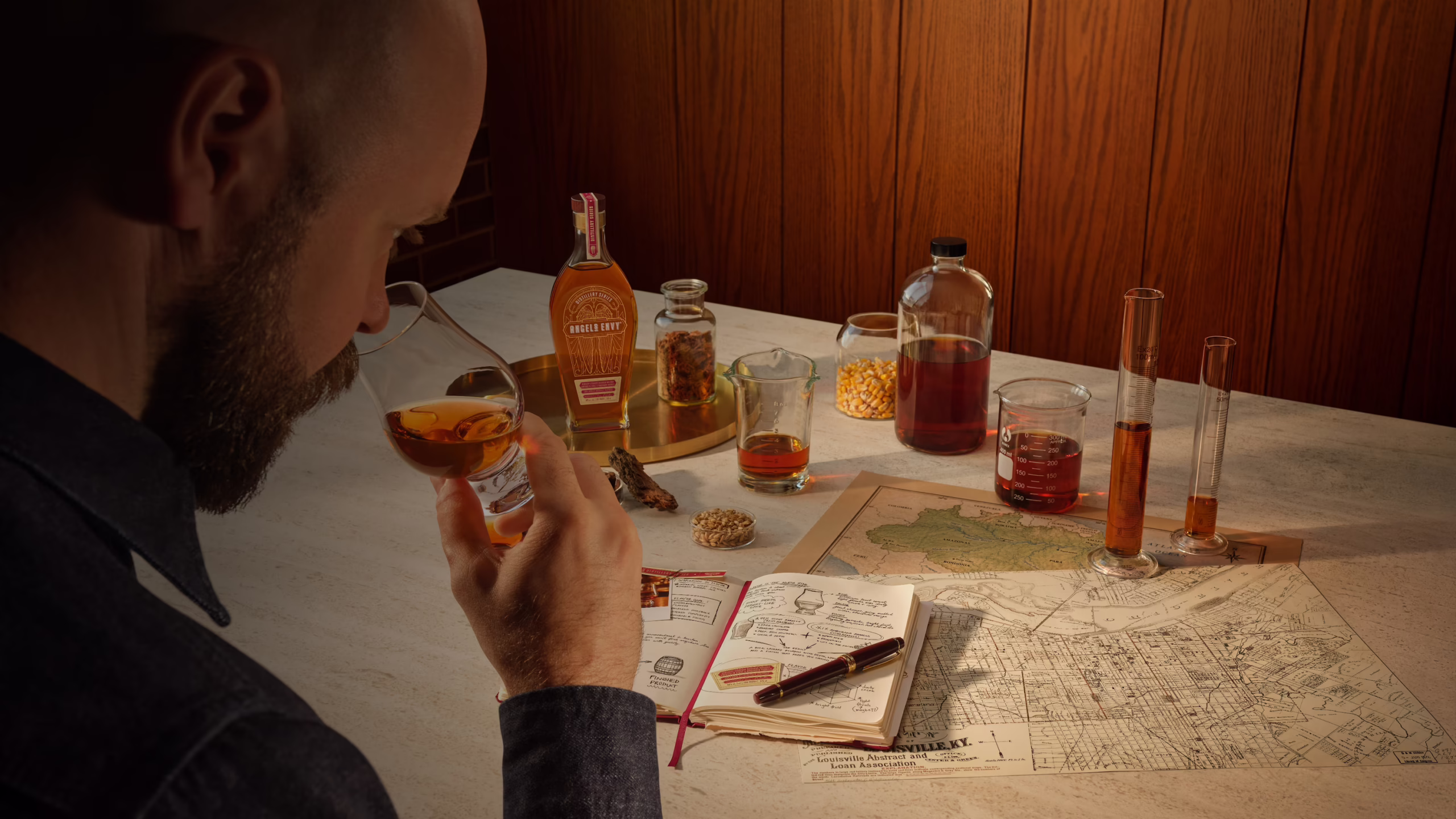 Master Distiller Owen Martin studying a glass of whiskey at a tasting table with bottles, beakers, a notebook, and a map of Louisville spread out in front of him.