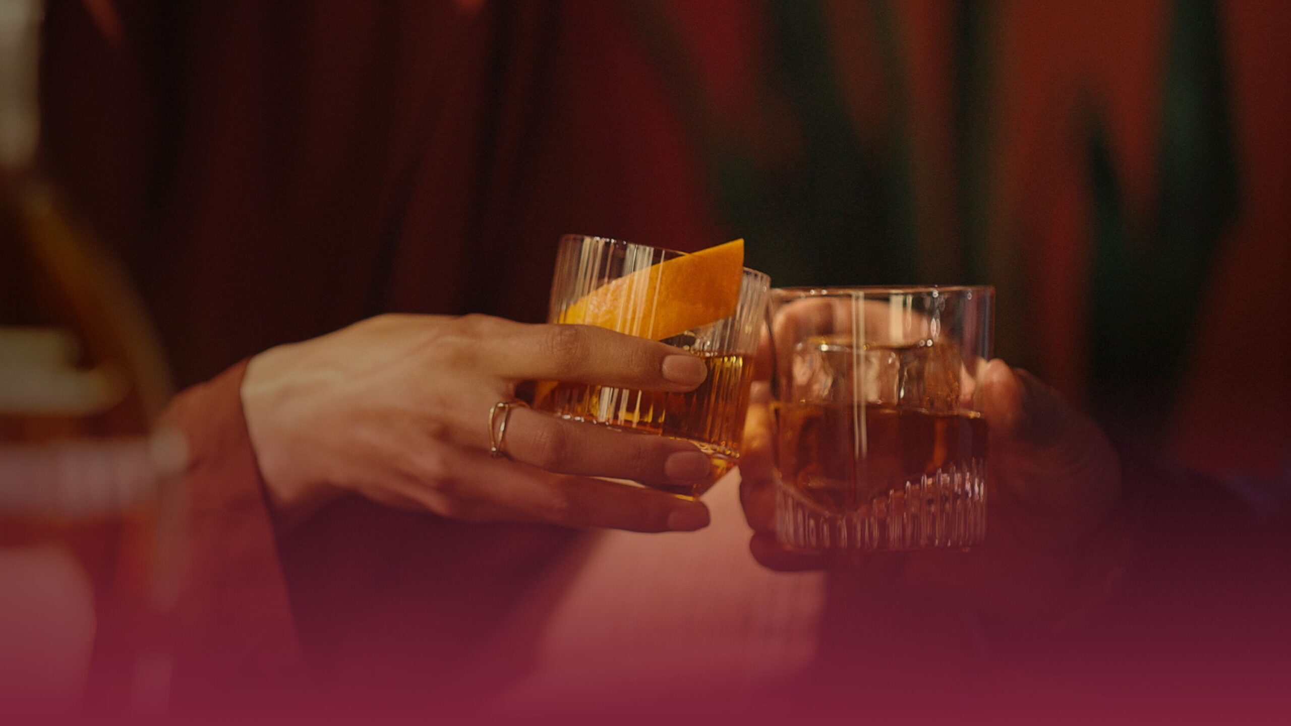 Two people clink whiskey glasses in a warmly lit setting. One glass holds an Old Fashioned garnished with an orange peel, the other contains a bourbon on the rocks.