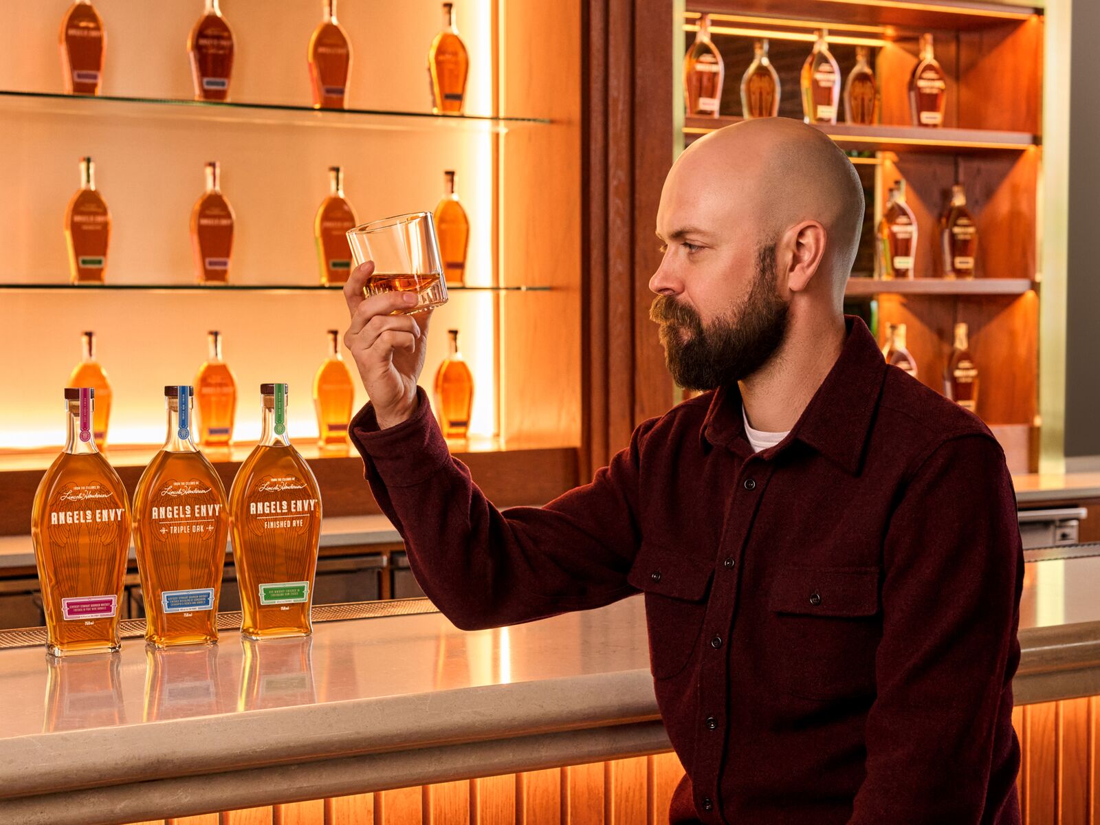 Master Distiller Owen Martin examining a glass of Angel’s Envy whiskey at a bar, with three Angel’s Envy bottles displayed beside him.