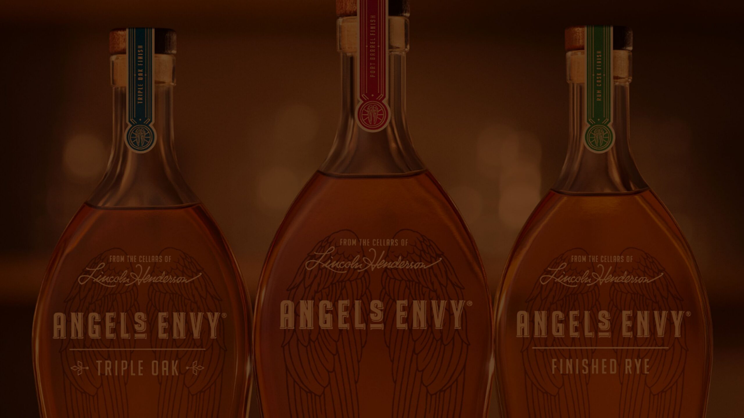 Close-up photo of three Angel’s Envy whiskey bottles, against a warm amber background.