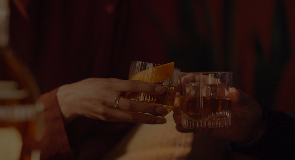 Two people clink whiskey glasses in a warmly lit setting. One glass holds an Old Fashioned garnished with an orange peel, the other contains a bourbon on the rocks.