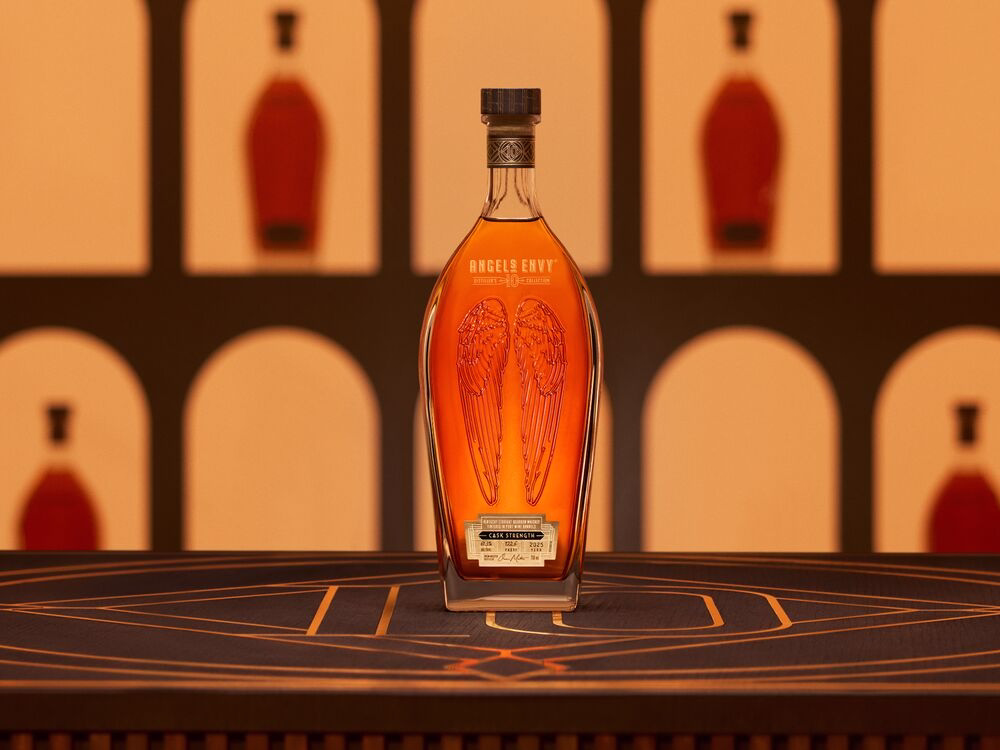 A bottle of Angel’s Envy 10 Cask Strength Bourbon displayed on a bar, with softly lit bottle silhouettes in the background.