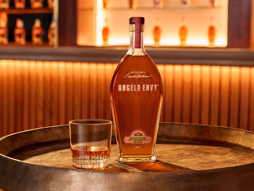 Angel’s Envy Whiskey displayed beside a whiskey neat poured in a rocks glass on a barrel-top table.