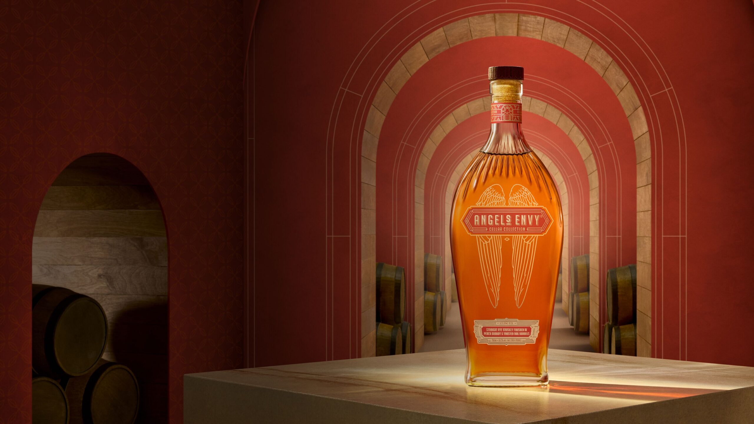 A bottle of Angel’s Envy Cellar Collection Rye displayed in a red corridor with oak arches and whiskey barrels visible in the background.
