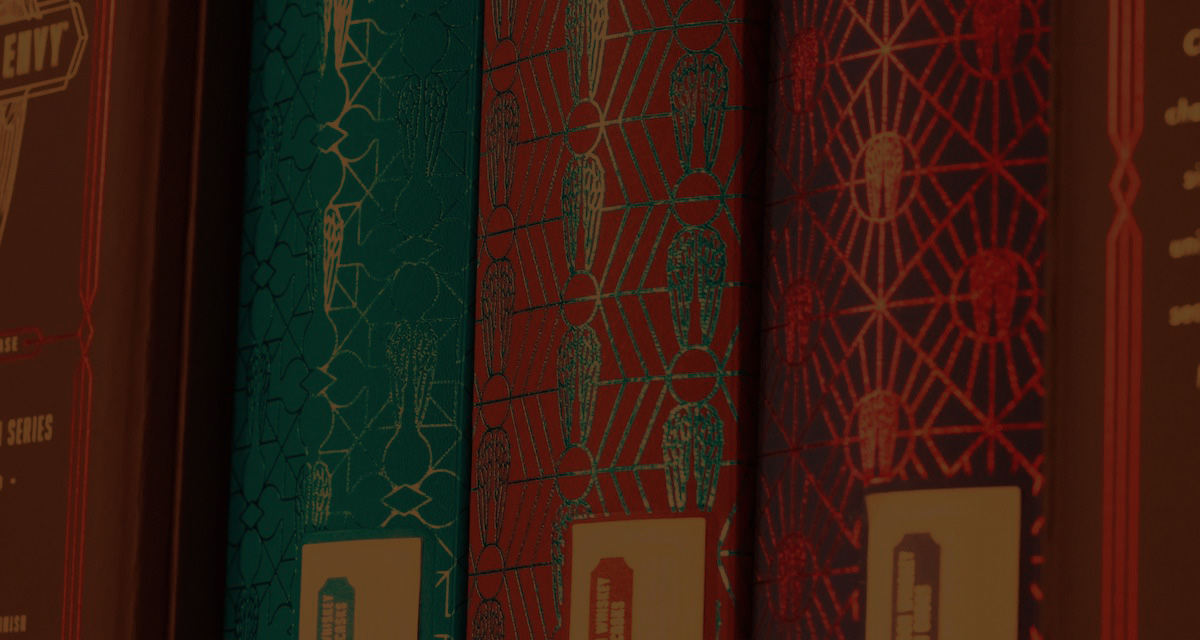 A close-up photo of the spines of decorative Angel’s Envy presentation boxes. In order, the three boxes are blue, red, and purple, each one featuring intricate geometric artwork incorporating the Angel’s Envy wings logo.