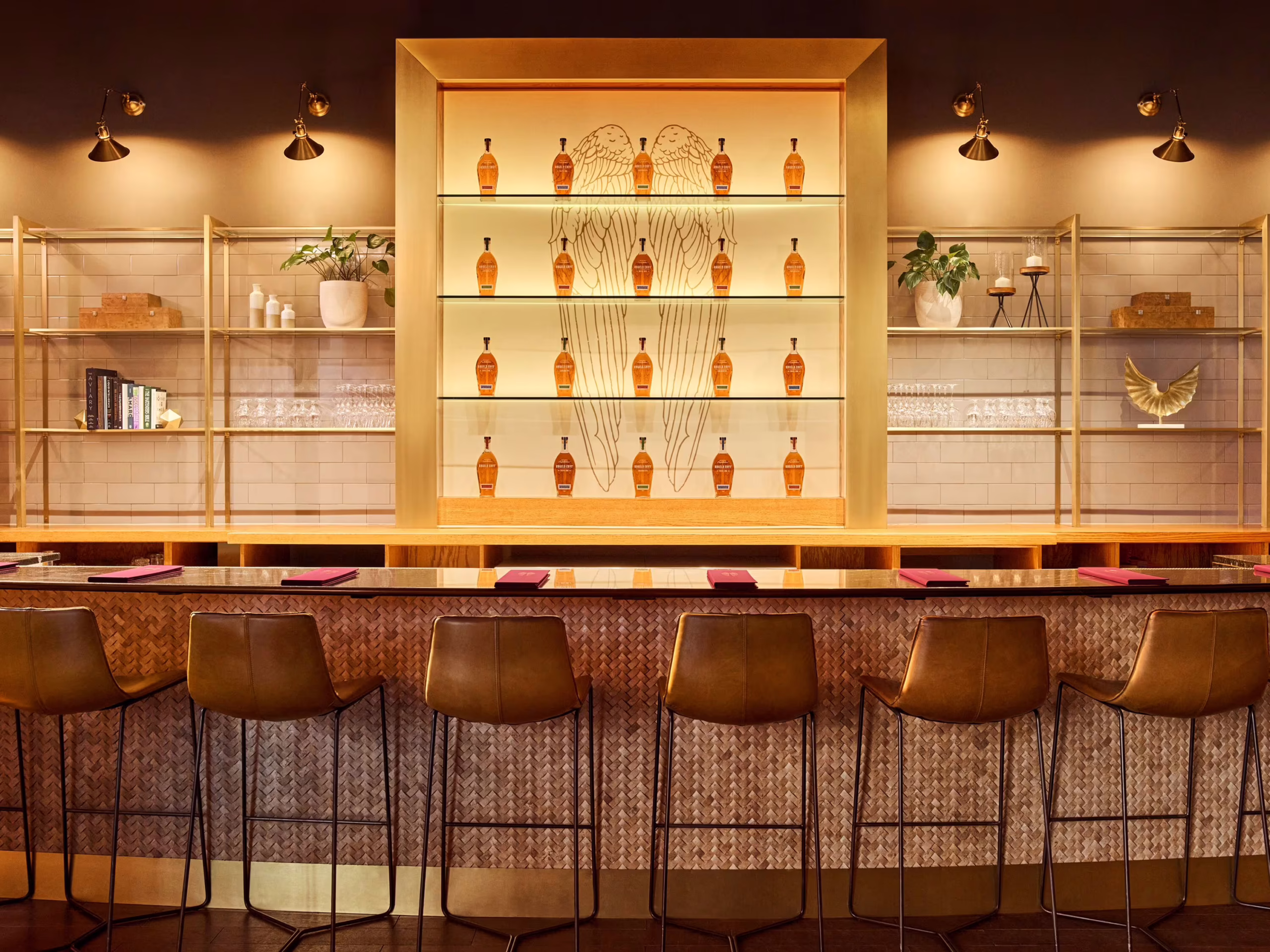 Image of the Angel’s Envy tasting bar with illuminated shelves of whiskey bottles arranged around a large wing illustration. Bar stools sit in the foreground in front of an inviting bar top.