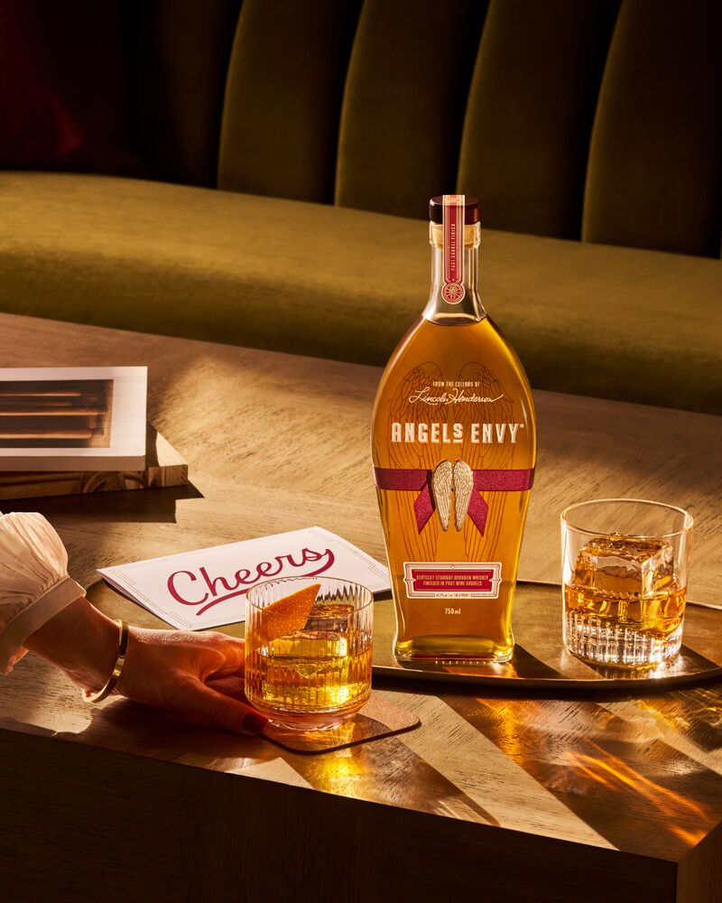 A bottle of Angel’s Envy on a brass serving tray, laying on a warmly lit table with two Old Fashioned cocktails, a handwritten ‘Cheers’ card, and a woman’s hand reaching for a glass.