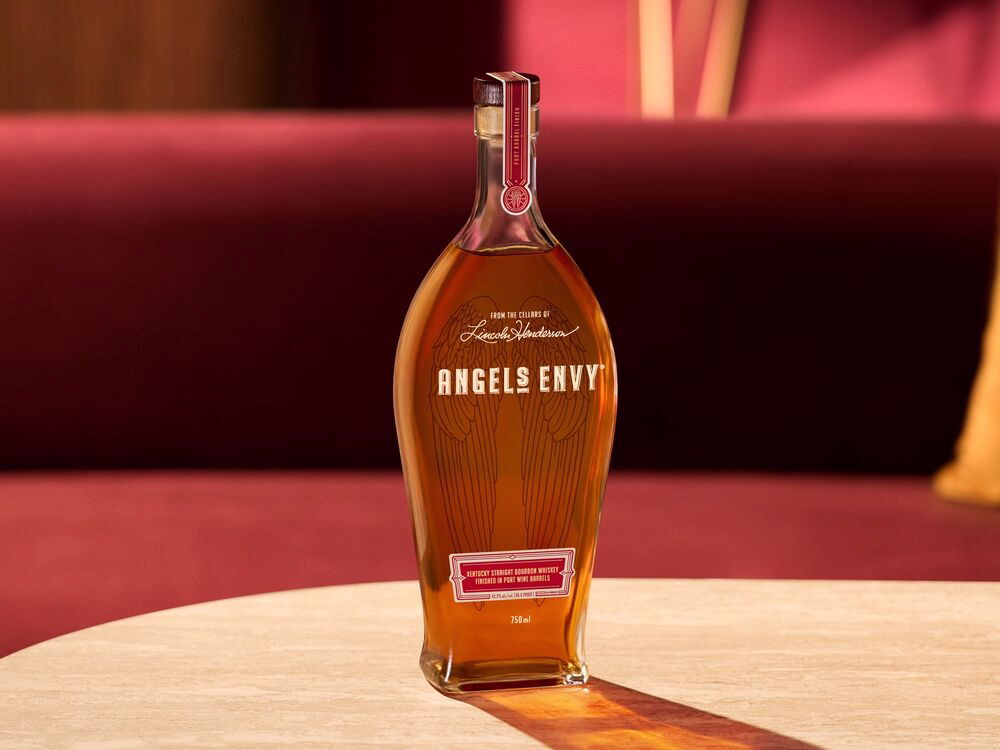 A bottle of Angel’s Envy Whiskey displayed on a wooden table, with red velvet furniture in the background.