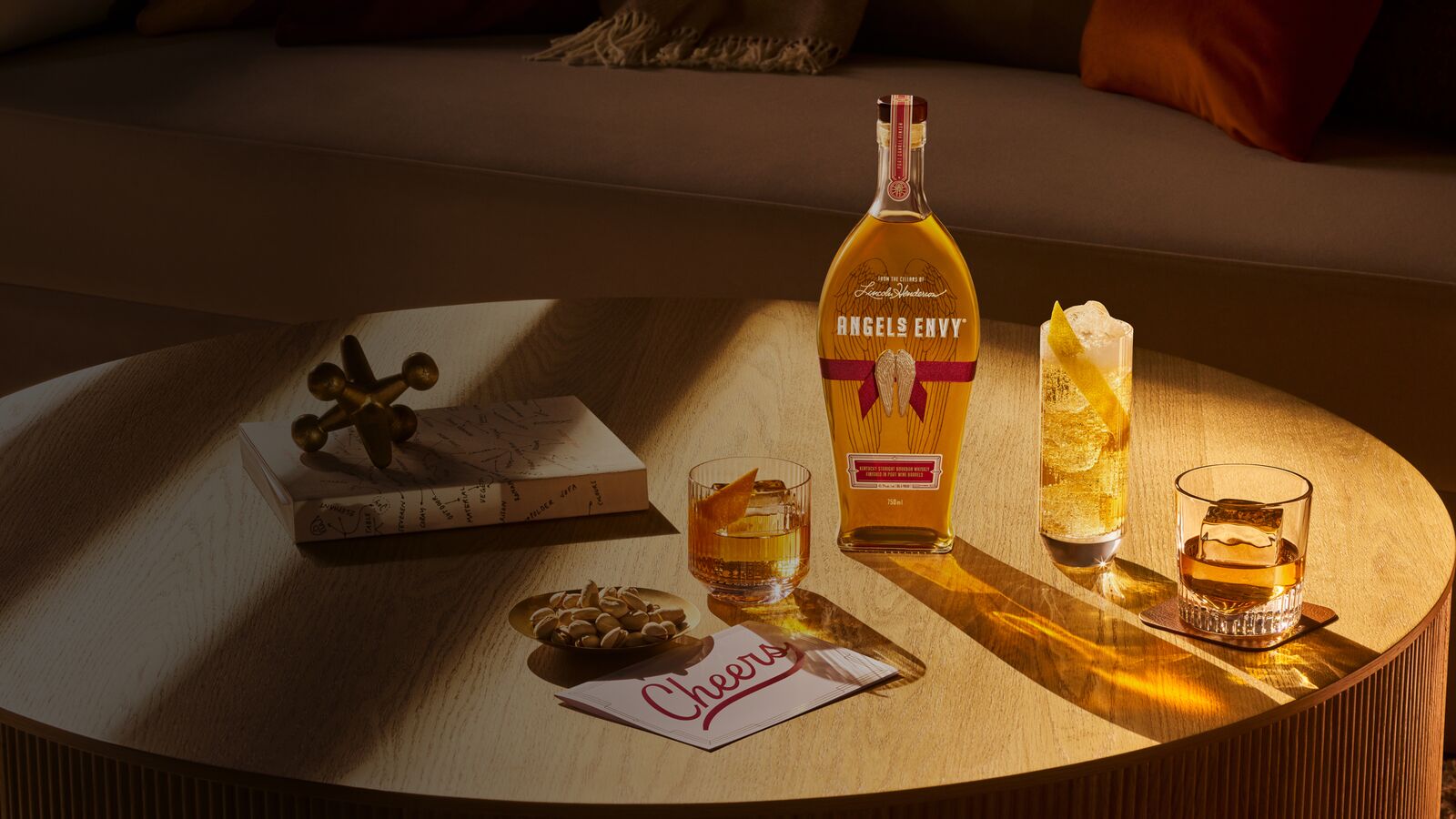 A ribbon-wrapped bottle of Angel’s Envy on a sunlit table next to three whiskey cocktails, a handwritten ‘Cheers’ card, a snack-sized bowl of pistachios, and table decor.