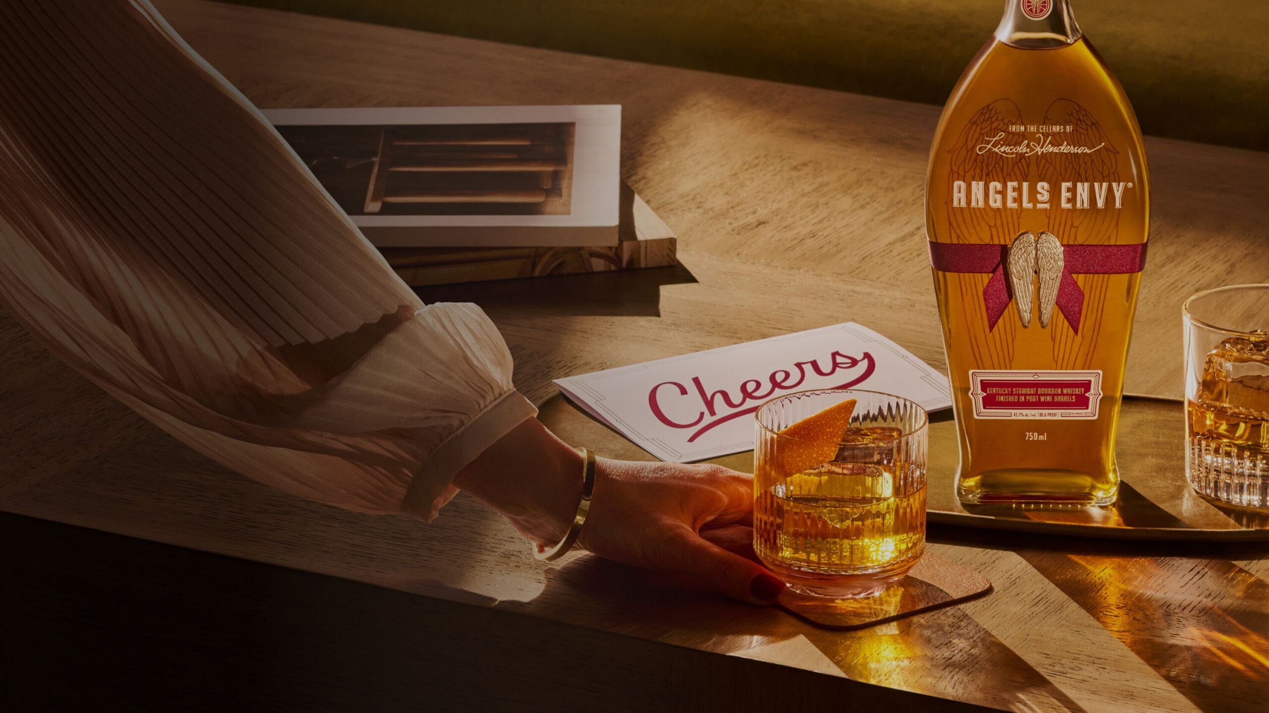 A bottle of Angel’s Envy on a brass serving tray, laying on a warmly lit table with two Old Fashioned cocktails, a handwritten ‘Cheers’ card, and a woman’s hand reaching for a glass.