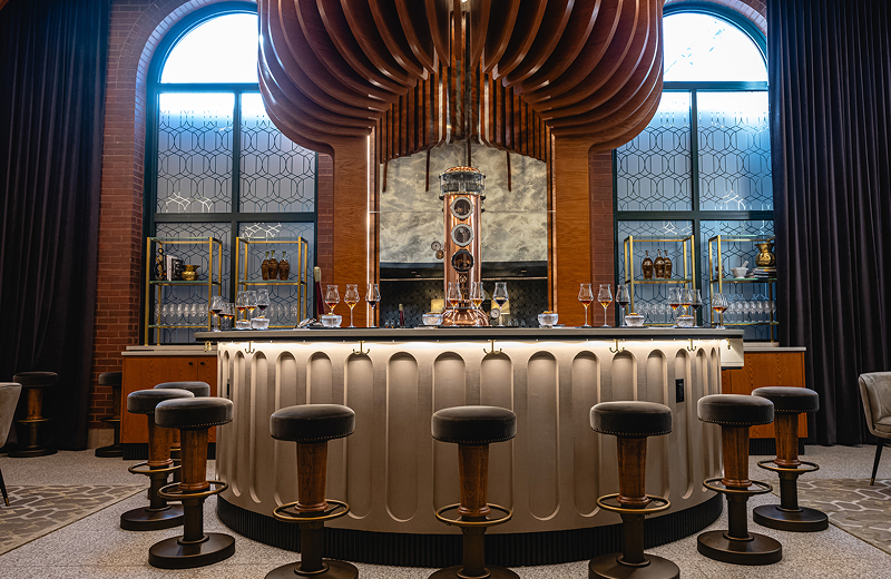 The elegant interior of Angel’s Envy Bar Ten/10 with arched windows, whiskey glassware, illuminated shelves, and a piece of custom equipment acting as a sculptural copper centerpiece.