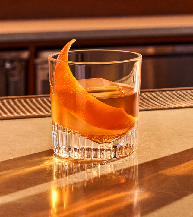 An Old Fashioned cocktail in a coupe glass garnished with an orange rind sitting on a clean granite bar.