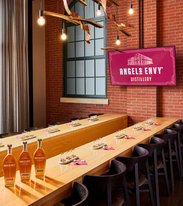 A tasting room at the Angel’s Envy Distillery with long wooden counters set with whiskey samples, three Angel’s Envy bottles, and a ruby display on a wall-mounted TV.