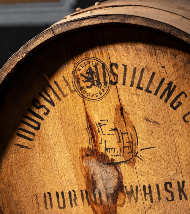 Close-up image of the textures on a wooden whiskey barrel stamped ‘Louisville’ and ‘Kelvin Cooperage,’ illuminated by warm sunlight.
