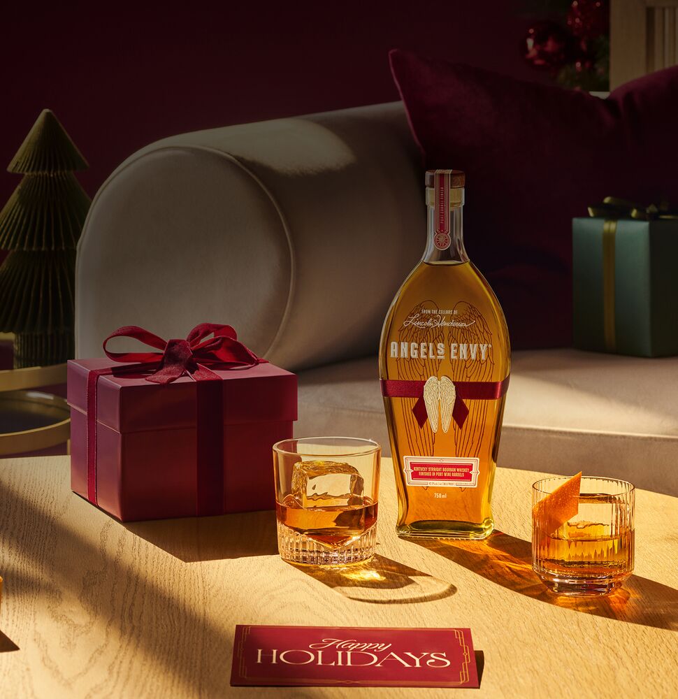 A bottle of Angel’s Envy Triple Oak Bourbon displayed next to three unique whiskey cocktails, a holiday card, and a gift on a warmly lit table.
