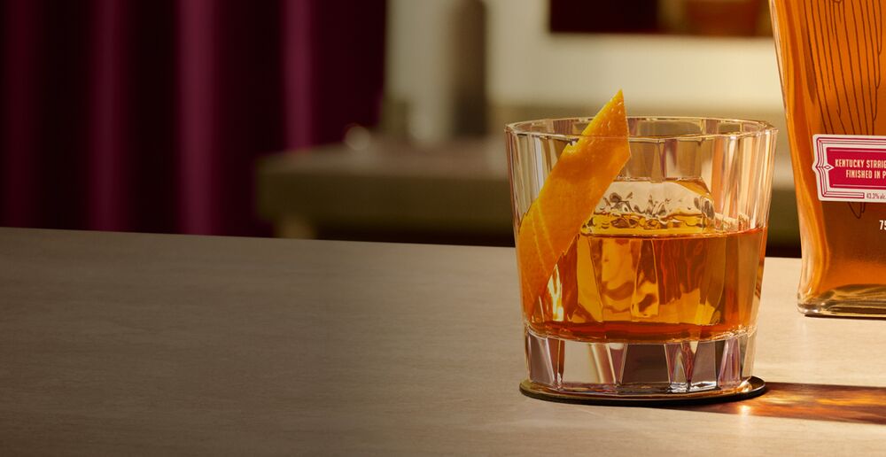 An Old Fashioned cocktail in a coupe glass, garnished with an orange rind. In the background, there’s a bottle of Angel’s Envy Port-Finished Bourbon.
