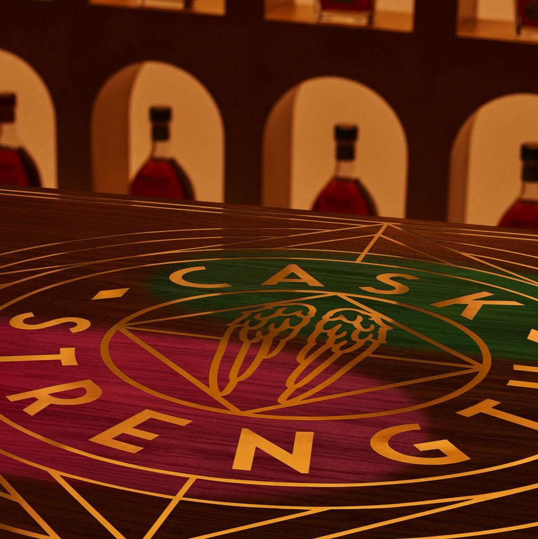 A teaser of the Angel’s Envy Spring 2026 Dual Cask Strength release within the Cask Archive room, featuring green and ruby spotlight casting across a logo-engraved tabletop.